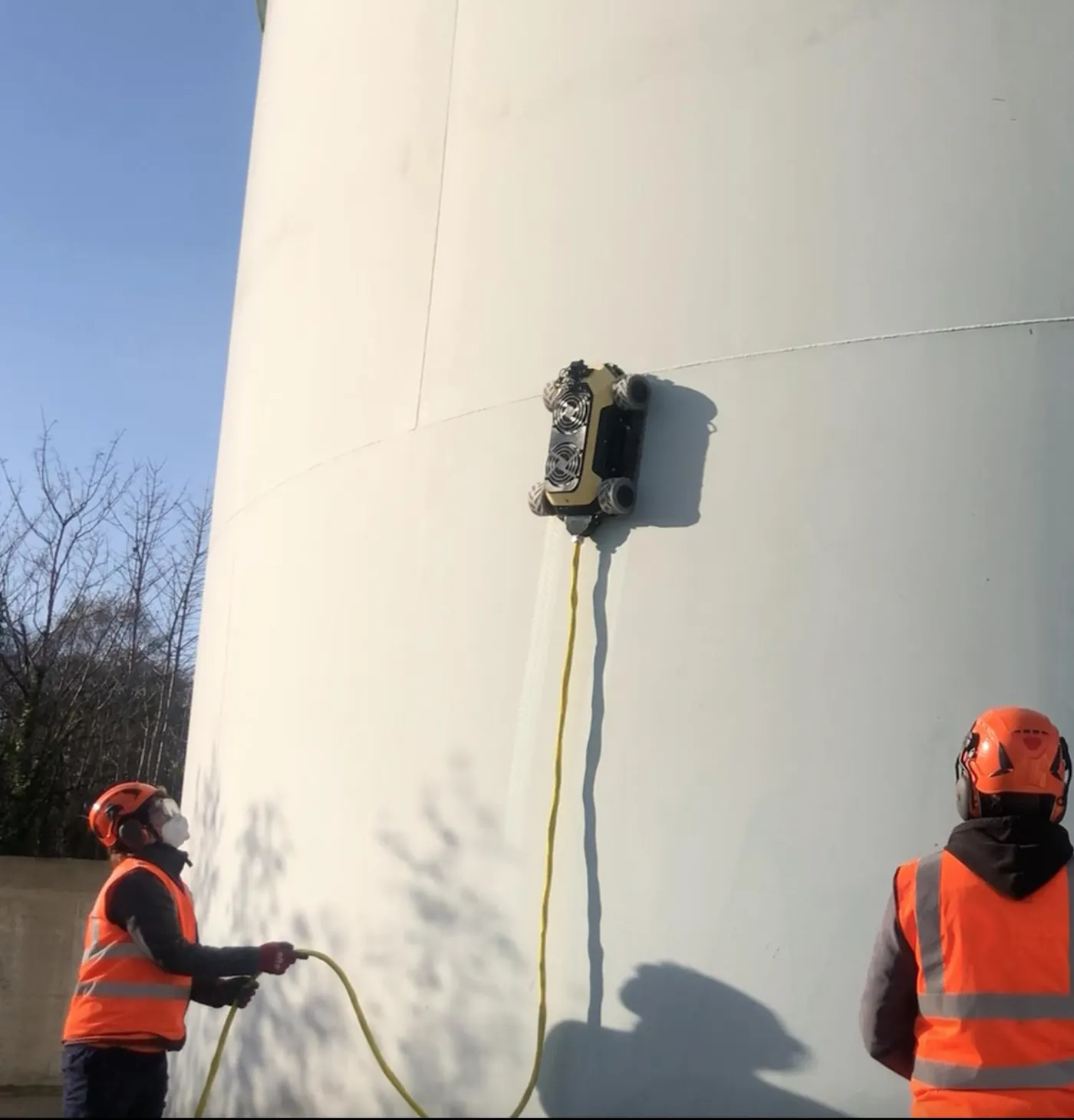 Robot fixé à la paroi verticale d’un grand réservoir blanc, deux opérateurs en casque et gilet orange le surveillent depuis le sol